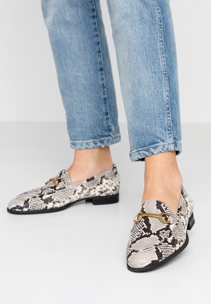 Fødder i lyseblå jeans iført hvide og sorte loafers med slangeskind og guldfarvet hestebid-detalje på en hvid gulv.
