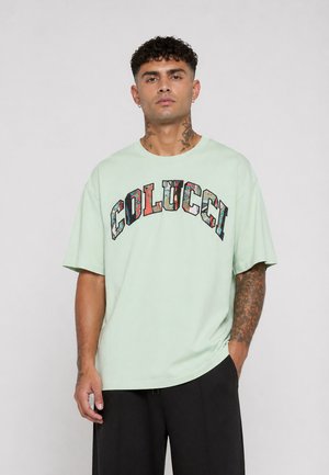 Mann med tatoveringer iført mintgrønn t-skjorte med fargerik "COLUCCI"-trykk og svarte bukser, stående mot en ensfarget lys bakgrunn.