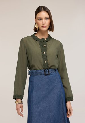 Kvinde med kort brunt hår iført olivengrøn bluse med bølgekant og guldknapper, blå højtaljet nederdel samt guldøreringe og armbånd.