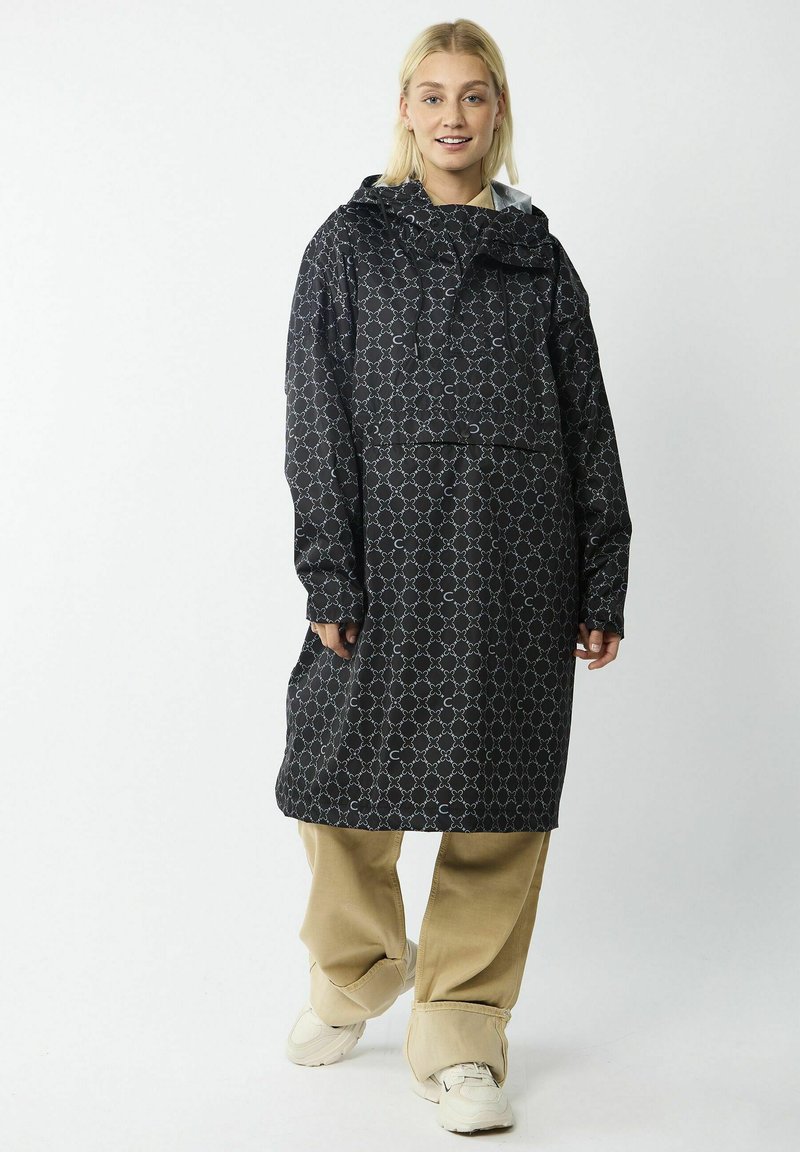 Zwarte regenjas met een geometrisch patroon in tinten van blauw. Voorzien van een capuchon, zijzakken en een relaxte pasvorm. Gekozen over beige broek en schoenen.