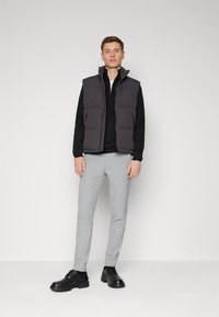 Gray padded vest over a black zip-up top, paired with light gray tailored trousers and black lace-up shoes. Simple, functional design.
