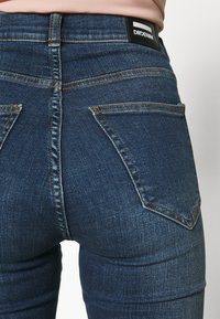 Jeans de ganga azul escuro com um design ajustado, apresentando um estilo clássico de cinco bolsos, costuras distintas e uma etiqueta de cintura com marca.
