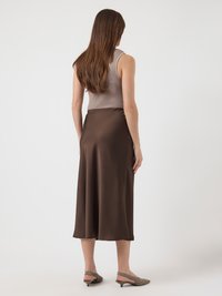 Mulher com cabelo castanho longo, vestindo uma blusa sem mangas em tom taupe, saia midi castanho escuro e sapatos de salto baixo em tom bege, de pé contra um fundo branco.
