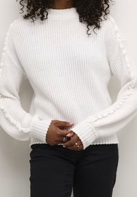 Witte gebreide trui met geribbelde manchetten en getextureerde zijaccenten. Voorzien van een hoge halslijn en een losse pasvorm. Gecombineerd met een zwarte broek.