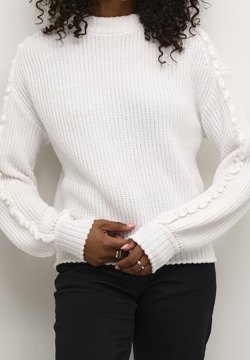 Witte gebreide trui met geribbelde manchetten en getextureerde zijaccenten. Voorzien van een hoge halslijn en een losse pasvorm. Gecombineerd met een zwarte broek.