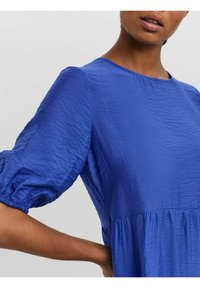 Modrá šaty s nafouklými rukávy a texturovaným povrchem, kulatým výstřihem a nařaseným pasem. Tkanina vypadá lehce a splývavě.