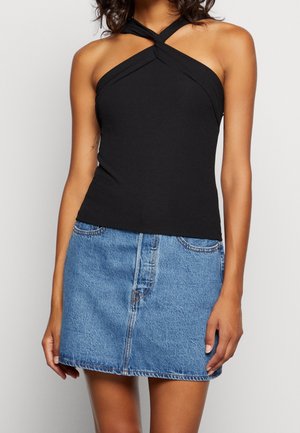 Vrouw draagt een zwarte haltertop met een gekruiste halslijn en een blauwe denim minirok, staand tegen een effen achtergrond.