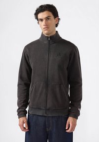 Hombre vistiendo una chaqueta de forro polar negra con cremallera y cuello alto, con bolsillos frontales, combinada con jeans azul oscuro, de pie frente a un fondo sencillo.