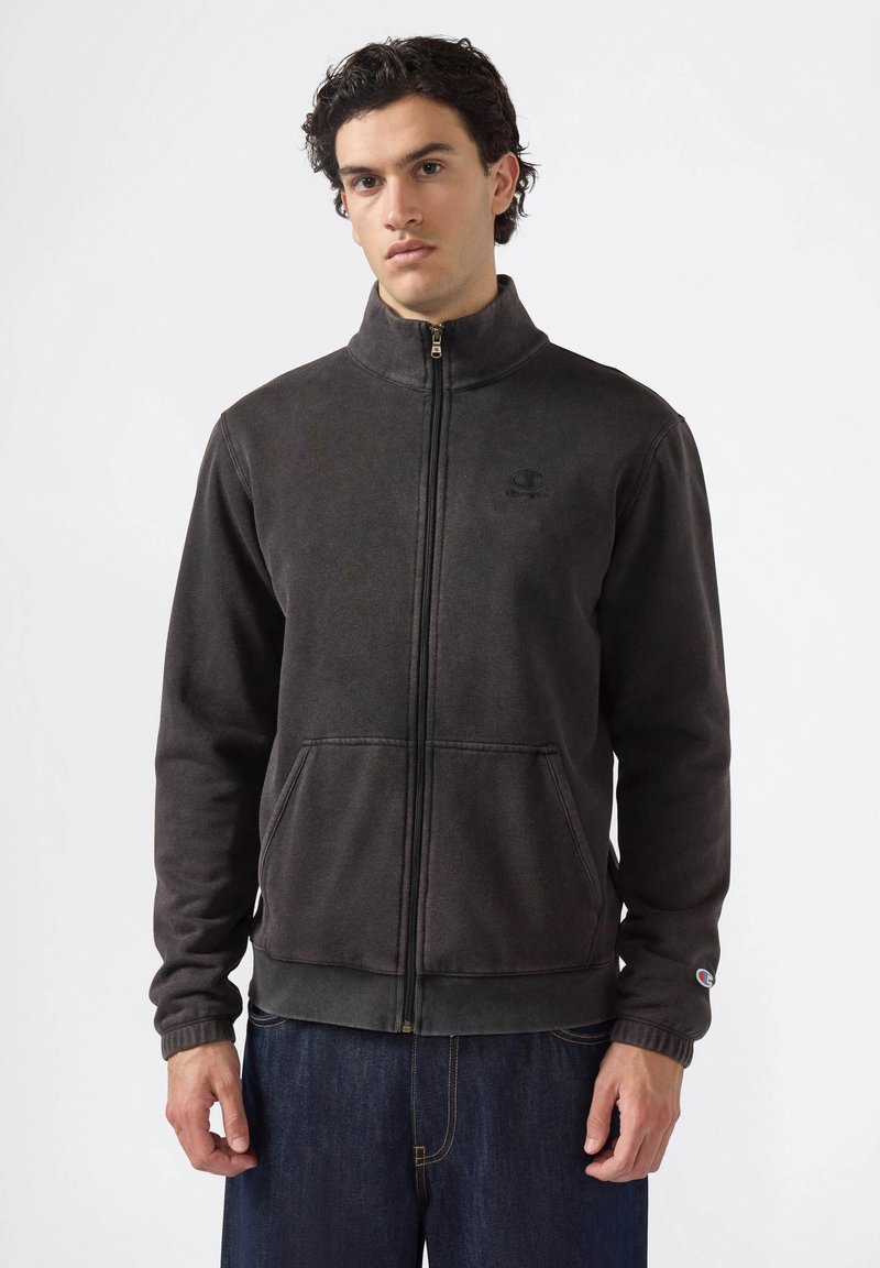 Hombre vistiendo una chaqueta de forro polar negra con cremallera y cuello alto, con bolsillos frontales, combinada con jeans azul oscuro, de pie frente a un fondo sencillo.