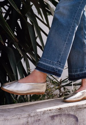 Scarpe slip-on metalliche oro con punta rotonda, texture morbida e suola naturale, abbinate a jeans blu con orlo sfilacciato.