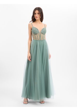 Frau in einem bodenlangen salbeigrünen Kleid mit transparentem, mit Perlen besetztem Oberteil und Spaghettiträgern, stehend vor einem schlichten weißen Hintergrund.