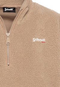 Beżowa bluza polarowa z półzamkiem, o fakturowanej powierzchni i wyszywanym logo "Schott" w kolorze białym z czerwonymi akcentami na lewej piersi.