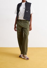 Černá péřová vesta přes bílý tričko, doplněná olivově zelenými kalhotami a vícebarevnými teniskami. Podlaha je jemně žlutá.