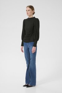Black long-sleeve blouse with gathered front detail, paired with flared blue jeans and pointed black shoes. Smooth fabric and structured design.