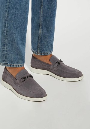 Gray suede slip-on shoes with a white rubber sole and decorative black hardware on the upper. The design features a smooth texture and rounded shape.