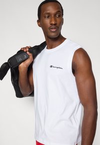 Λευκό βαμβακερό αμάνικο πουκάμισο με μαύρο λογότυπο "Champion" στο στήθος, κρατημένο πάνω στον ώμο με μαύρη τσάντα με λουρί.