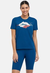 T-shirt blu a maniche corte con grafica di montagne in bianco, rosso e teal. Abbinato a pantaloni corti da bicicletta blu, texture liscia e design casual.