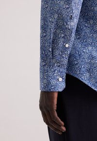 Una mano dalla pelle scura accanto a un polsino di camicia a maniche lunghe con motivo paisley blu e bottoni bianchi, su uno sfondo chiaro e neutro.