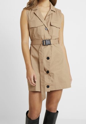 Robe beige sans manches à boutons, avec poches poitrine à rabat, taille cintrée par une ceinture avec boucle noire, portée avec des bottes noires montant jusqu'aux genoux.