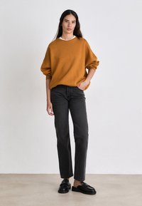 Suéter oversized mostaza, pantalones vaqueros negros de pierna recta y zapatos negros sin cordones; tejido texturizado con puños acanalados y una camiseta blanca debajo.