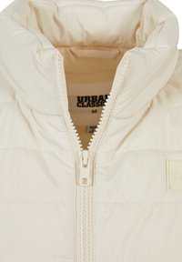 Pehme beespruun jope kõrge kraega, ees tõmblukuga ja väikese logomärgiga. Siledat tekstuuri ja horisontaalsete õmblustega.