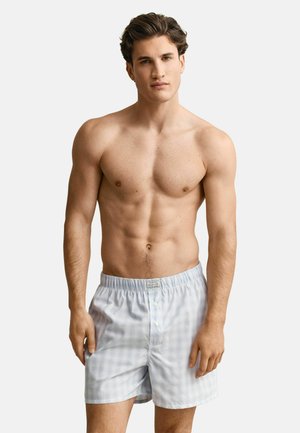 Jonge man met kort bruin haar, gekleed in lichtblauw geruite boxershorts, staand tegen een effen witte achtergrond.