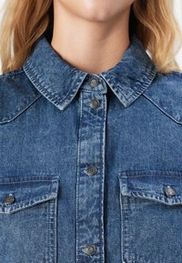 Close-up of a person wearing a blue denim jacket with a collar, front buttons, and two chest pockets with flaps and buttons.
