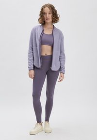 Lila Fleecejacke mit Reißverschluss, kombiniert mit einem passenden Sport-BH und Leggings. Das Model trägt weiße Sneaker. Insgesamt glatte Textur.