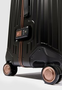 Schwarzer Hartschalenkoffer mit vertikalen Rillen, bronzefarbenen Akzenten, eingebautem Schloss und vier Rollen mit glänzendem Bronzelack.