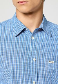 Camisa de cuadros azul y blanca con cuello abotonado, presenta botones blancos y un pequeño logo de Wrangler en el bolsillo izquierdo.