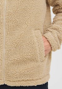 Chaqueta de forro polar beige con una superficie texturizada, con cremallera frontal y bolsillos laterales. Los puños y el dobladillo están acabados de manera pulcra.