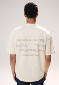 Cremfarbendes Baumwoll-T-Shirt mit kurzen Ärmeln. Mit großen, kontrastierenden Drucktexten auf dem Rücken, einschließlich Markenname und Slogans.