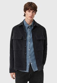 Tmavě šedá texturovaná košilová bunda s límečkem, předními kapsami a zapínáním na knoflíky, přepážkovaná přes modrou džínovou košili.