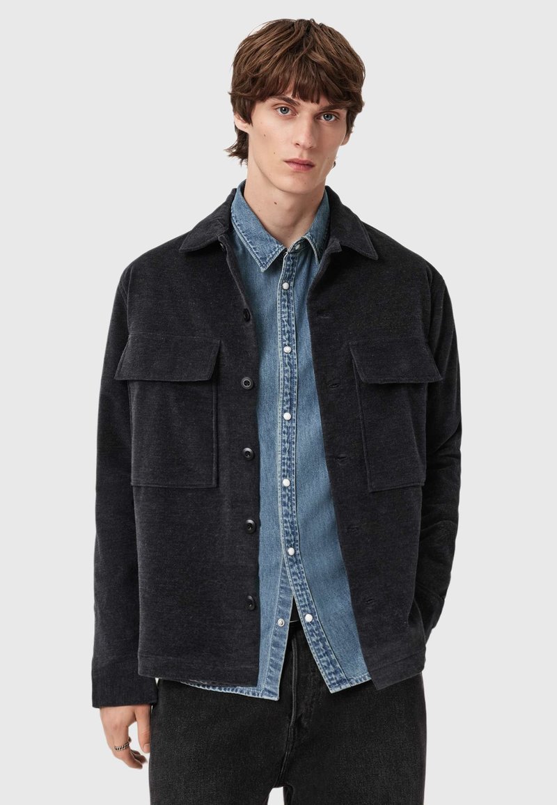 Tmavě šedá texturovaná košilová bunda s límečkem, předními kapsami a zapínáním na knoflíky, přepážkovaná přes modrou džínovou košili.