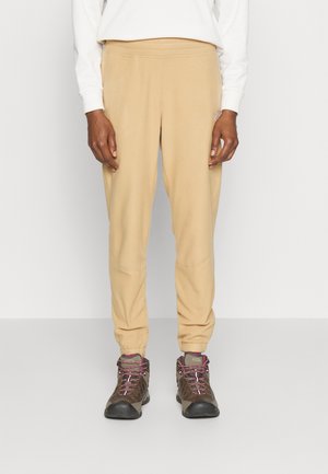 Persona con pantalones de chándal beige y botas de senderismo marrones, de pie frente a un fondo liso.