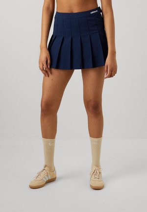 Kobieta w granatowej plisowanej spódnicy Adidas, beżowych skarpetach do połowy łydki i beżowych sneakersach Adidas stoi na tle jednolitego tła.