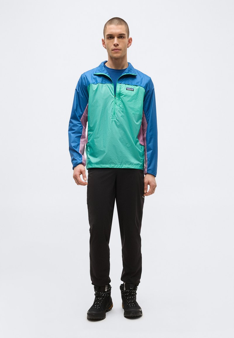 Junger Mann, der frontal steht und eine blau-grün-lila Windjacke, schwarze Hose und schwarze Wanderschuhe auf einem einfarbigen Hintergrund trägt.