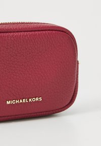 Červená koženková kosmetická taštička s texturovaným povrchem a zlatým kováním. Má zaoblené hrany a zipové uzavření. Na přední straně je logo "MICHAEL KORS".