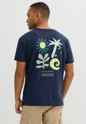 Hombre con camiseta azul marino con diseño en la espalda que incluye un sol tropical amarillo y verde, una palmera, una hoja, una ola y el texto "Verano Infinito".