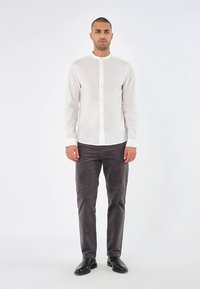 Hvid skjorte med knapper og ståkrave, båret med grå bukser. Glat stof, tætsiddende design, let glans og lange ærmer.