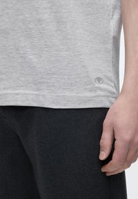 Κοντινό πλάνο ενός ατόμου που φοράει ανοιχτό γκρι μπλουζάκι και σκούρο παντελόνι, με το δεξί χέρι χαλαρό στο πλάι.