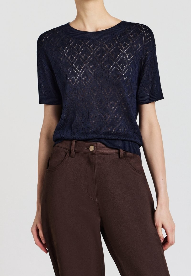 Femme portant un haut en maille bleu marine à manches courtes avec motif en losanges et un pantalon marron taille haute avec poches avant et fermeture à boutons.