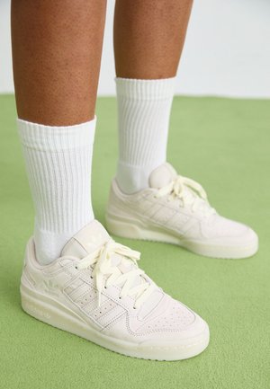 Ben met witte geribbelde sokken en crèmekleurige Adidas sneakers staand op een lichtgroene ondergrond.
