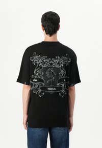 Black cotton t-shirt featuring a large white graphic on the back, including ornate designs and the word "HUGO" within a banner.