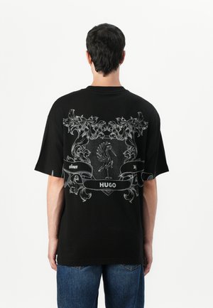 T-shirt di cotone nera con una grande grafica bianca sul retro, che include design ornamentali e la parola "HUGO" all'interno di un banner.