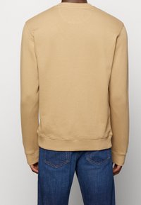 Felpa khaki con maniche lunghe, polsini e orlo a coste, scollatura rotonda e tessuto morbido al tatto. Indossata con jeans in denim blu.