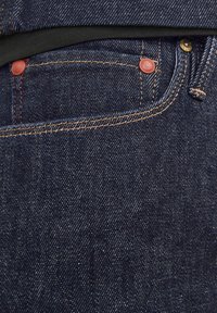 Vaqueros de mezclilla azul oscuro con costuras en contraste, que presentan bolsillos remachados en cobre y un acabado texturizado. Incluye una cinturilla negra en la parte superior.