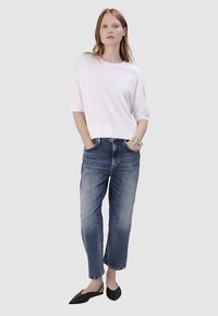 Weißes Baumwoll-T-Shirt mit kurzen Ärmeln, kombiniert mit blauen Jeans aus Denim. Die Jeans haben eine lockere Passform und einen ausgefransten Saum. Schwarze flache Schuhe vervollständigen das Outfit.