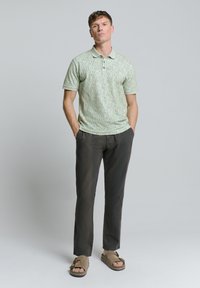 Groene geprinte polo met kraag, gecombineerd met donkere broeken en bruine sandalen met gesps. Model staat met handen in de zakken.