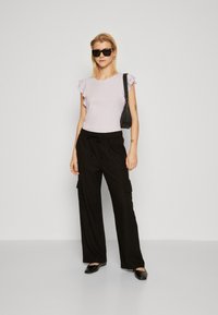 Haut lilas à manches volantes associé à un pantalon cargo noir à jambes larges avec poches latérales. Le mannequin porte des lunettes de soleil noires et des chaussures.